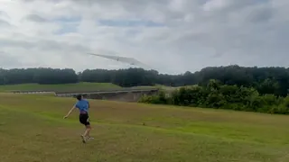 A person runs and hand-launches a large white flying wing across a grassy field under an overcast sky